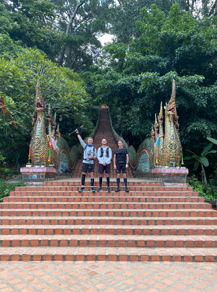 How To Run a 100 Miles Ultramarathon (And How I Did It) 3 3 men in sports gear pose outside of a stairwell to a temple in Chiang Mai, Thailand.