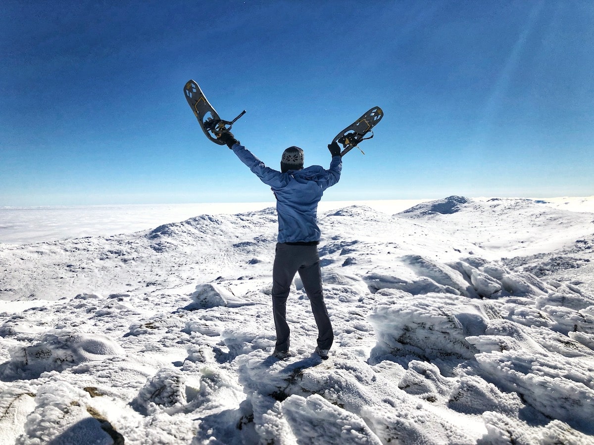 Climbing Mount Kosciuszko in Winter | Summiting Mount Kosciuszko in winter Hiker raises his snowshoes above his head on top of a snow-capped mountain in Australia