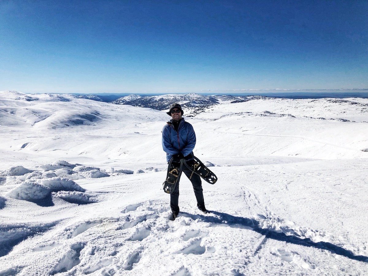 Climbing Mount Kosciuszko in Winter | climbing mount Kosciuszko independently Man holding ski shoes on top of Mount Kosciuszko, Australia