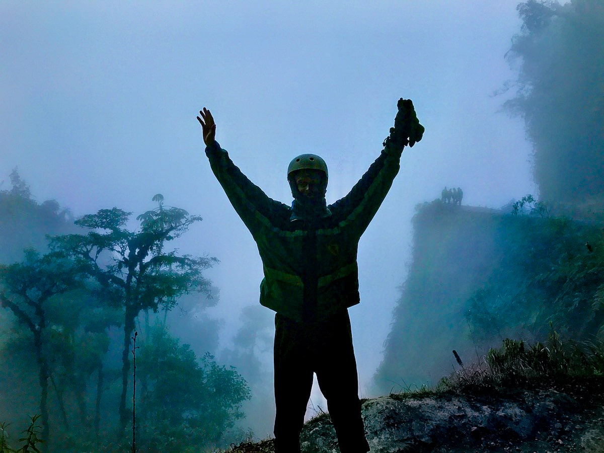 Cycling Down Bolivia's Death Road (How Not To Die!) | death road bolivia tourist A male tourist with hands aloft in the air as others cycle down Bolivia's Death Road in the misty background.