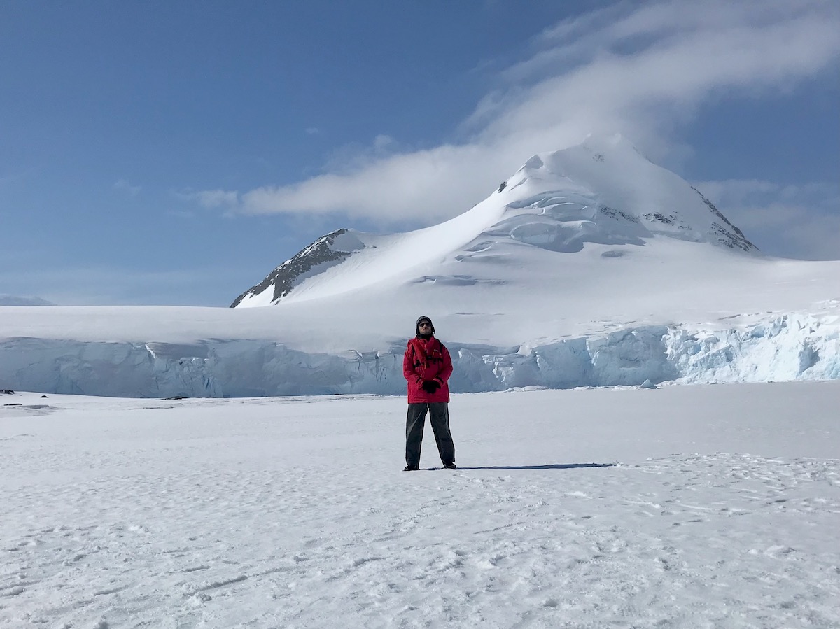 What To Expect on Your First Trip To Antarctica | Antarctica gear Man in red parka and grey hiking pants stands in the snowy landscape of Antarctica