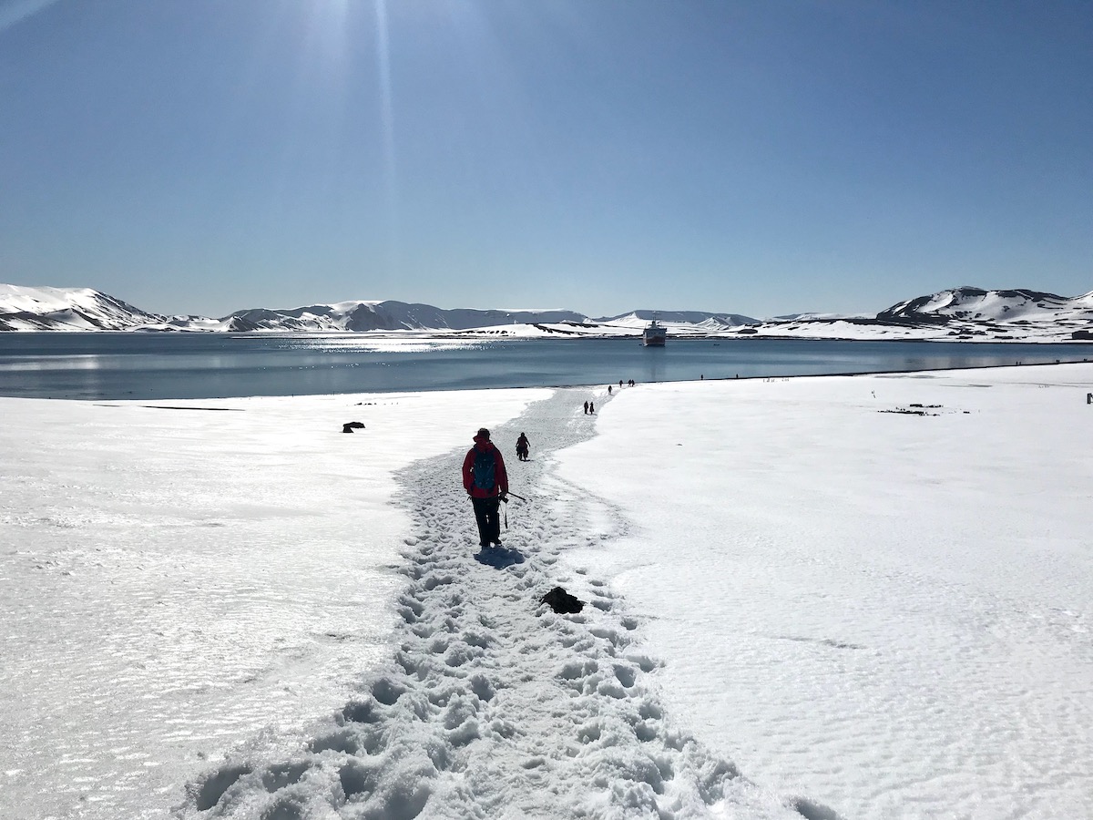 What To Expect on Your First Trip To Antarctica | Antarctica trail People individually walk on a trail in the snow towards a blue lake during a trip to Antarctica