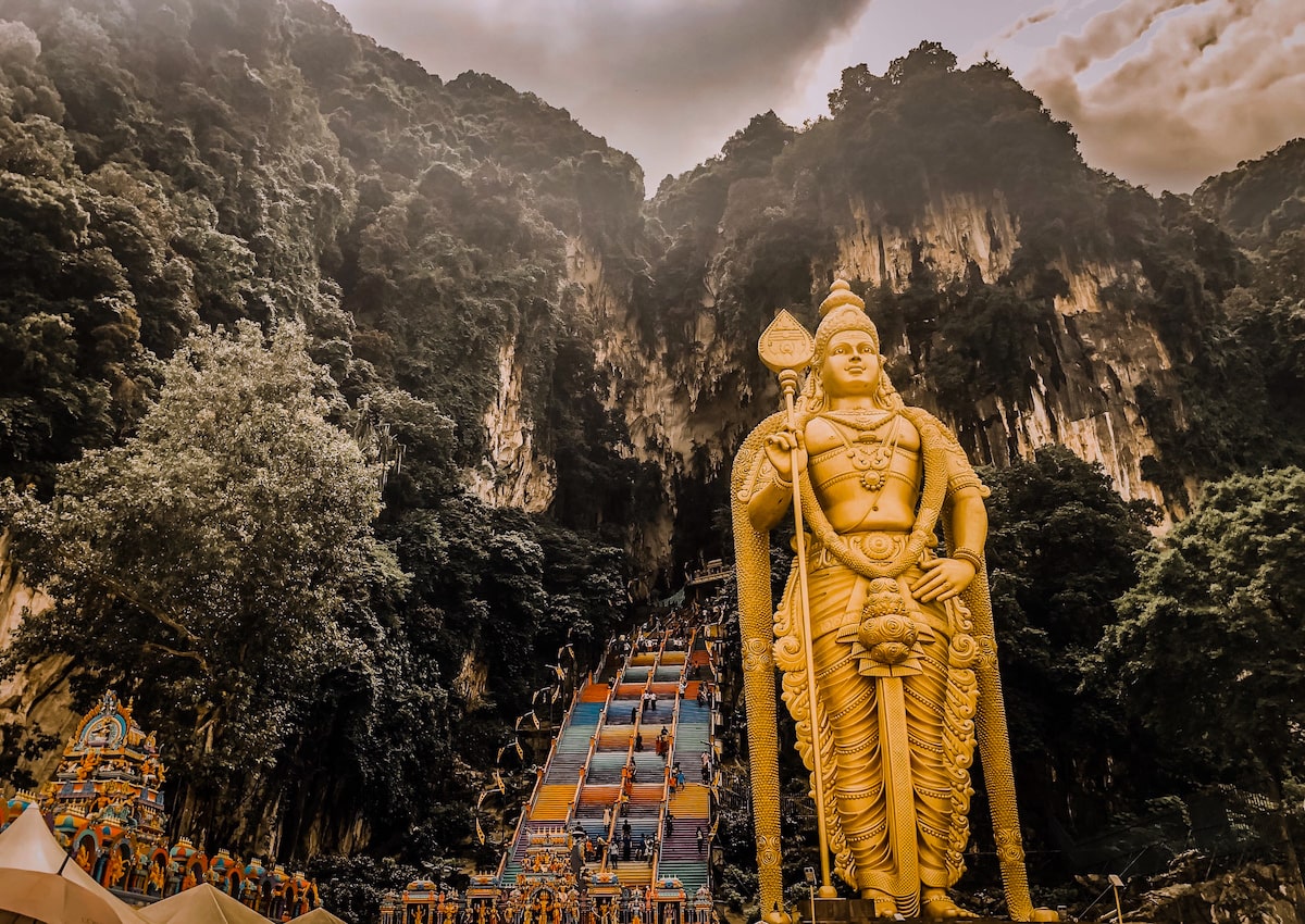 Thaipusam Festival at Batu Caves in Kuala Lumpur 2025 | Batu Caves Kuala Lumpur Malaysia A large, majestic golden Lord Murugan stands tall in front of multicoloured steps and large limestone caves in Kuala Lumpur, Malaysia