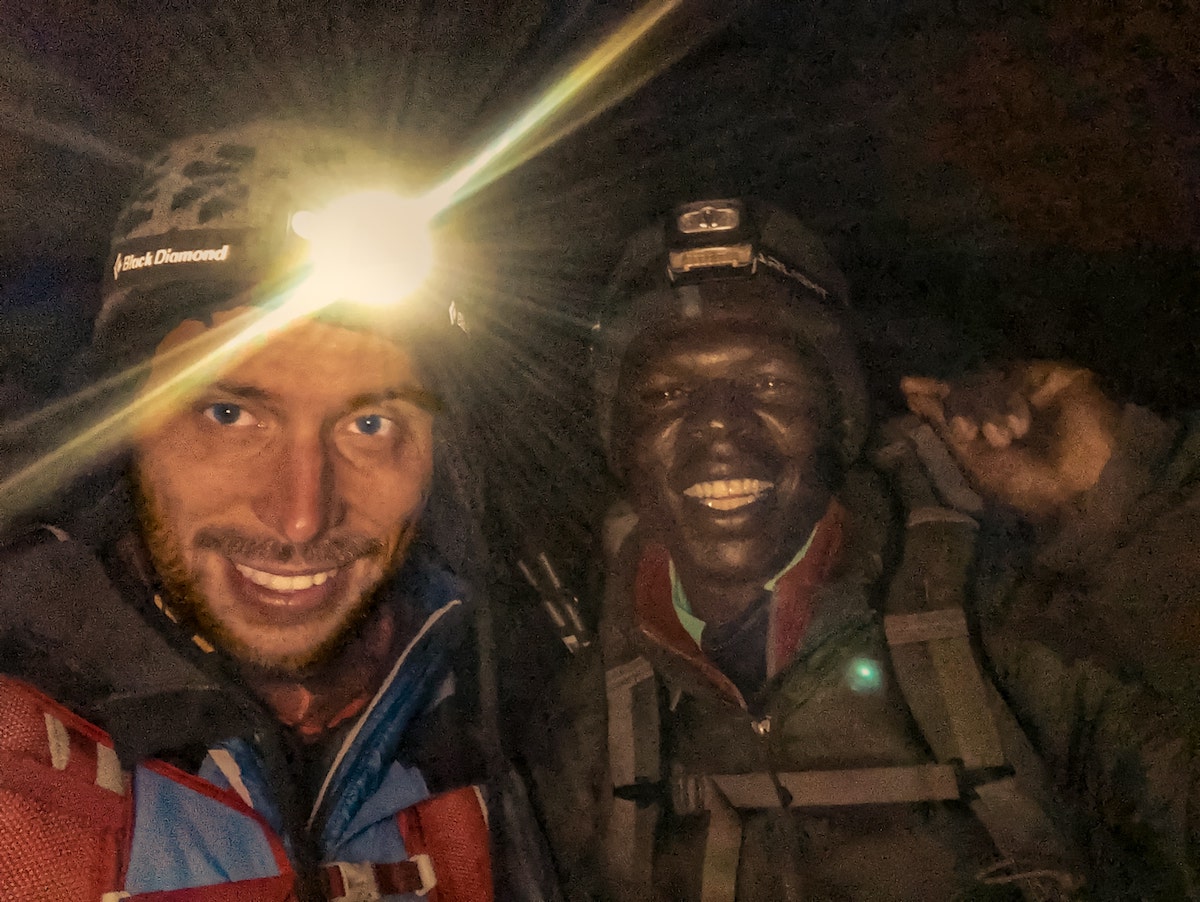 The Beginner's Guide To Climbing Mount Kilimanjaro | Climbing Kilimanjaro in dark Two men smile at the camera up close with head torches on their foreheads.