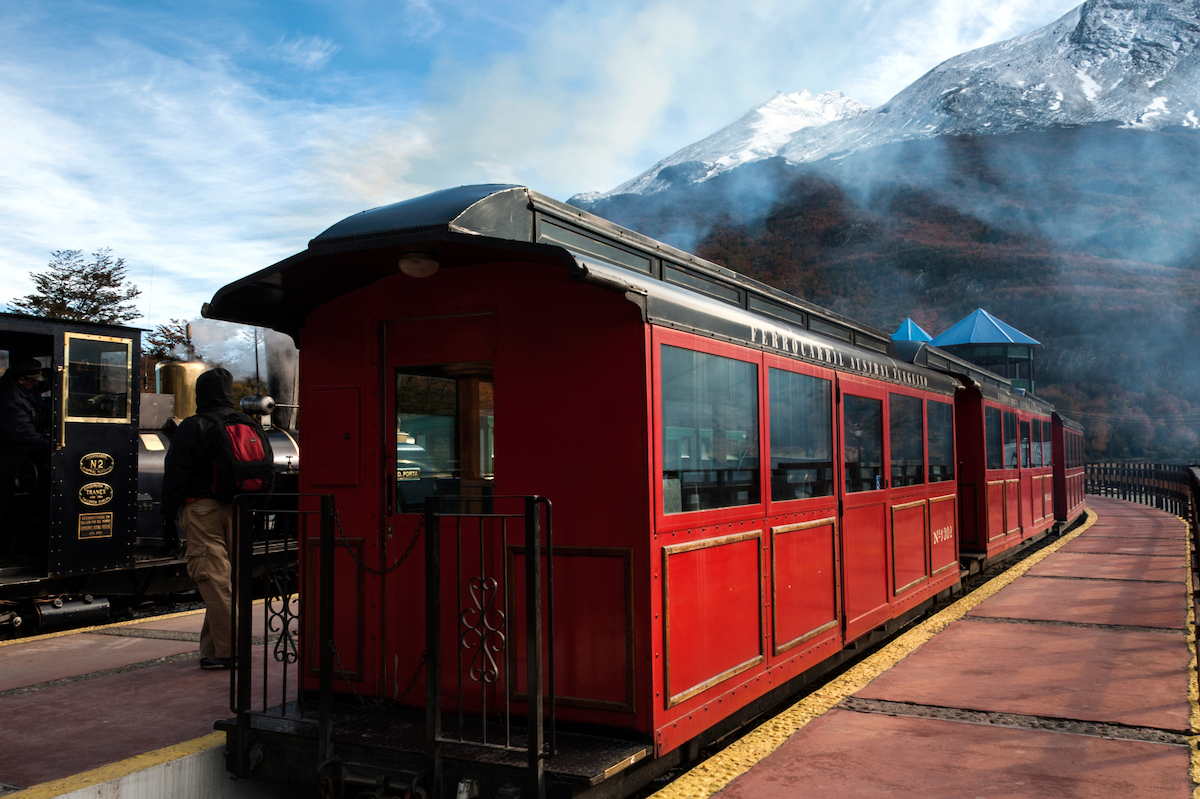 23 Best Things To Do in Ushuaia, Argentina | End of World Train Tierra del Fuego A red tram stops alongside a snow-capped mountain in Tierra del Fuego, Ushuaia, Argentina