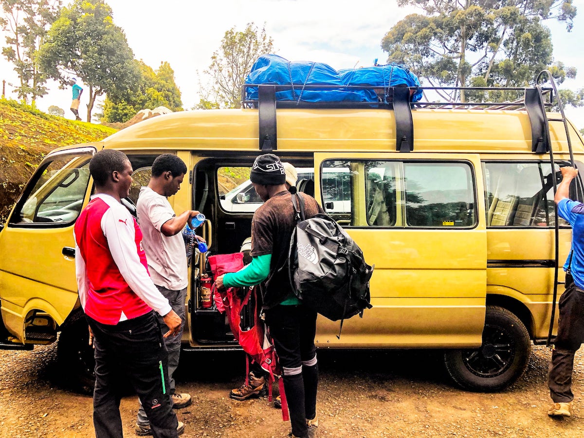 The Beginner's Guide To Climbing Mount Kilimanjaro | Entrance to Kilimanjaro A team of climbers wait outside of a yellow van.