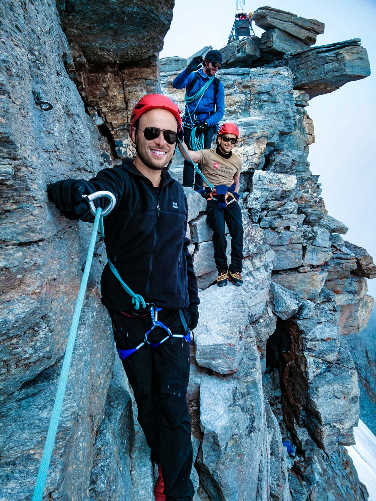 Mont Blanc Climb (The Beginner's Guide To Summiting in 2024) | Gran Paradiso rocks Three men pose for a smile on a mountain with jagged rocks, all of them roped in to one another.