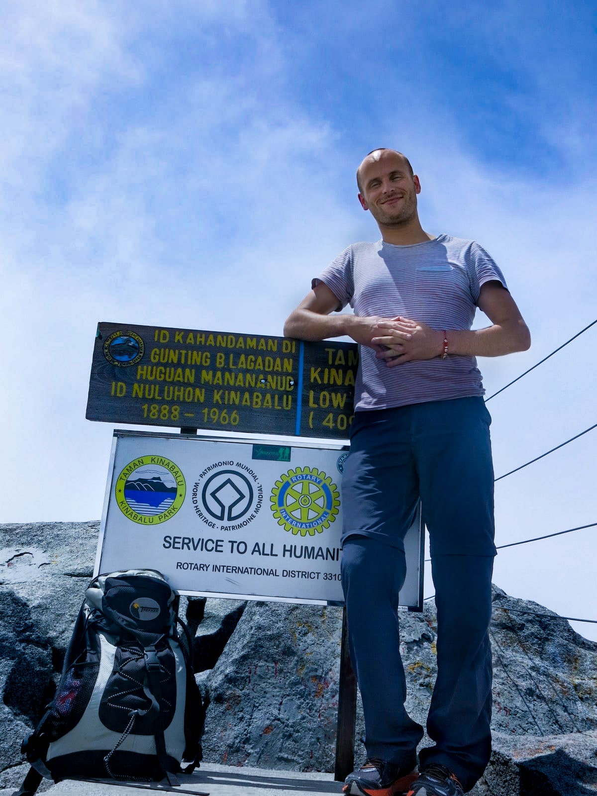 Climbing Mount Kinabalu Tips & Info For First-TimersĀ | Hiking Mount Kinabalu A man smiles as he leans on a sign with Bahasar writing on the summit of Mount Kinabalu, in Malaysian Borneo.