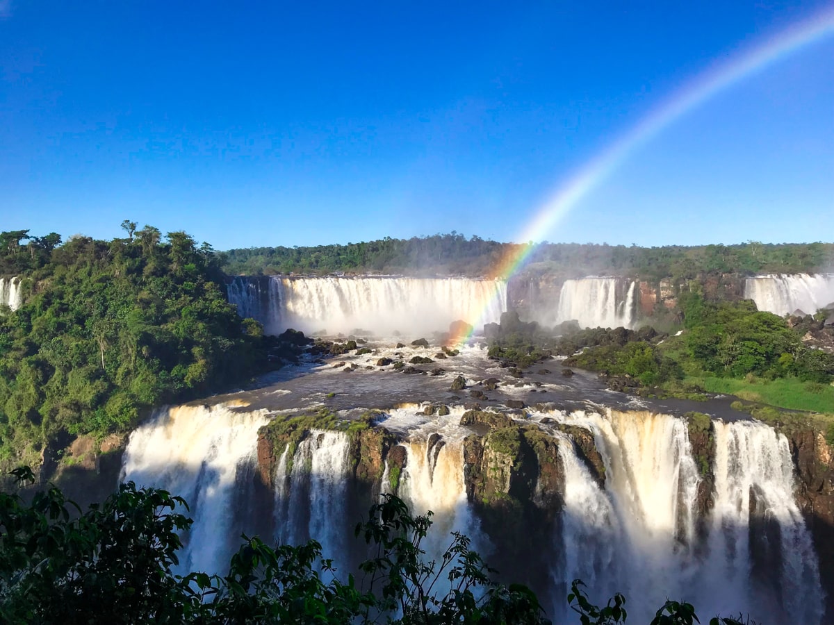 The Ultimate Guide To Visiting Both Sides of Iguazu Falls | Iguazu Falls Brazil A rainbow shines over a slew of waterfalls on a sunny day from The Iguazu Falls Brazil side