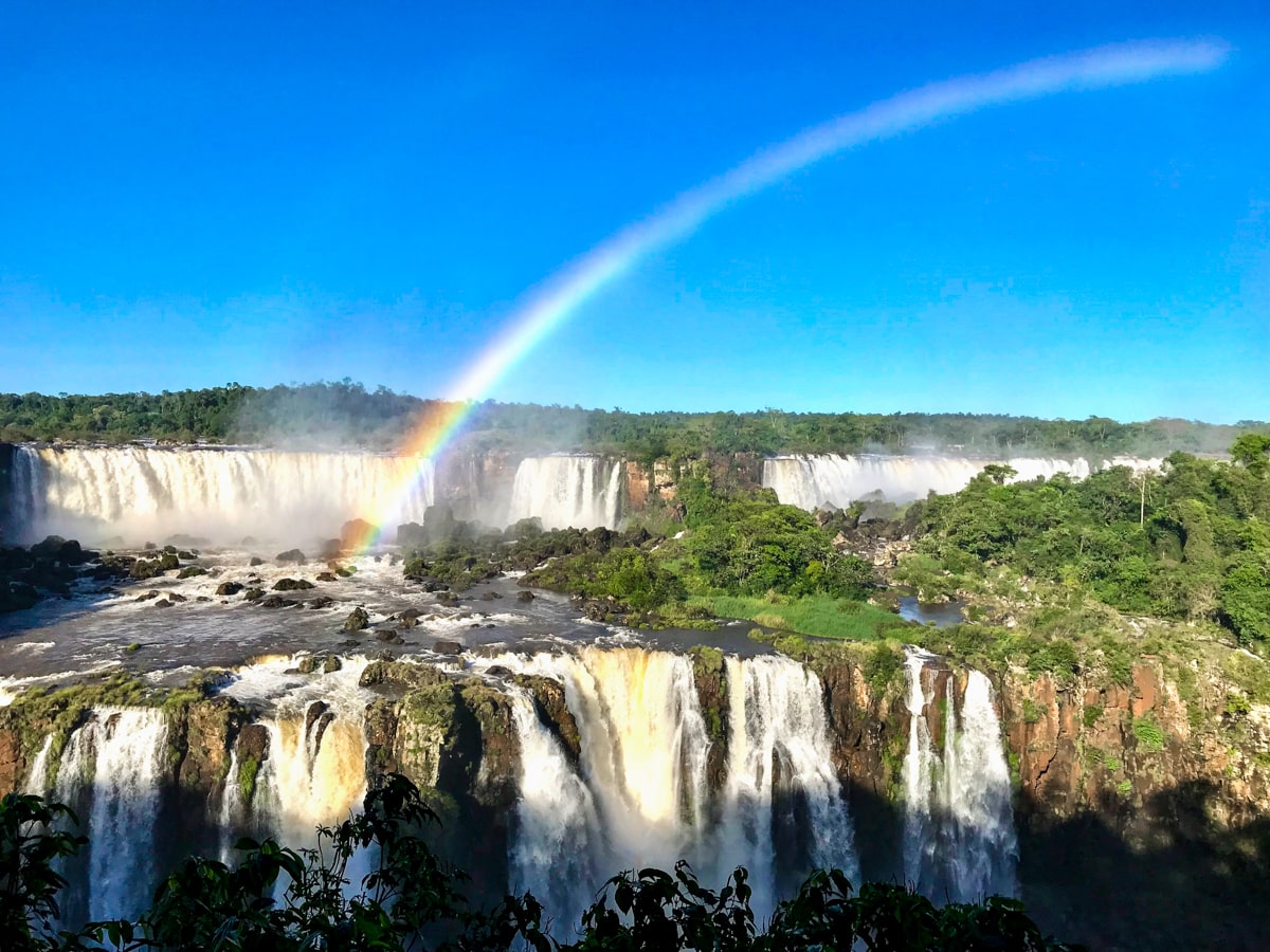 The Ultimate Guide To Visiting Both Sides of Iguazu Falls | Iguazu Falls with rainbow A rainbow shines over a gigantic waterfall, giving a treat to tourists visiting Iguazu Falls