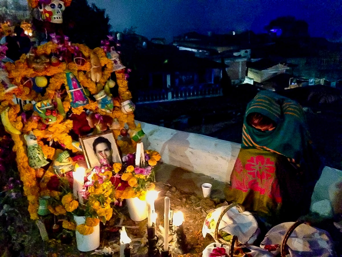16 Famous Landmarks in Mexico (& How To See Them in 2024!) | Janitzio day of the dead A lady wrapped in a scarf lowers her head next to a decorated grave, with a photo of a young man.