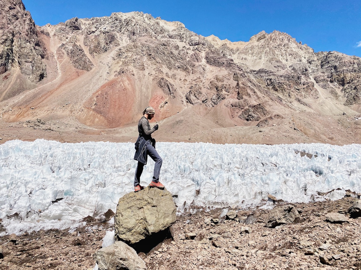 How Hard is it To Climb Aconcagua? | Las Heras Aconcagua 1 A mountain climber checks his sunglasses on Aconcagua while standing on a rock near glacier ice, with brown mountain landscapes and a bright blue sky behind him