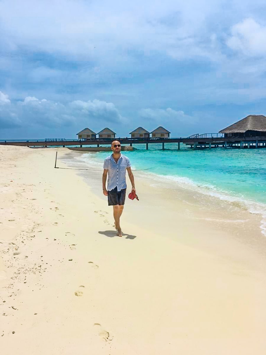 How To Plan a Trip To The Maldives | Maldives beach A man in a white shirt and blue shorts walks along the beach in a Maldives resort