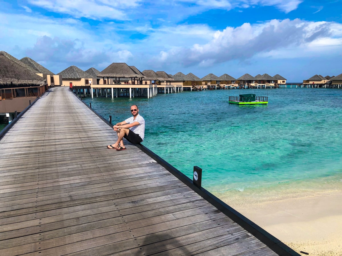 How To Plan a Trip To The Maldives | Maldives tourist A tourist in the Maldives sits on a wooden walkway with the backdrop of water villas and beautiful blue sea