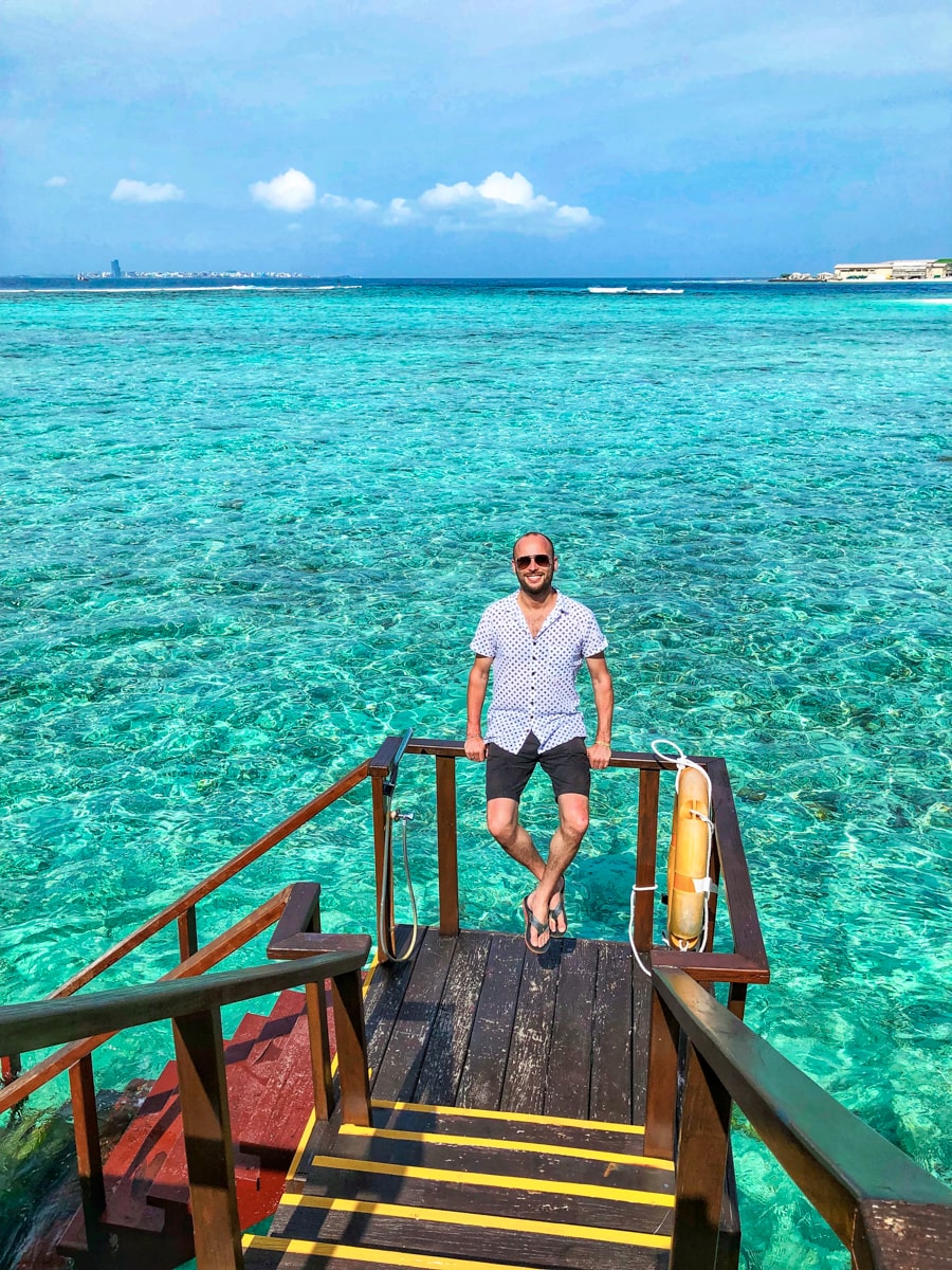 How To Plan a Trip To The Maldives | Maldives view from bungalow A male tourist with sunglasses smiles on a balcony with the clear blue sea in Maldives