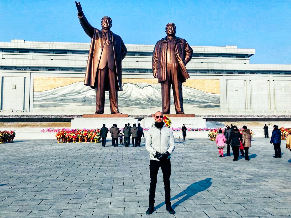 SafetyWing Review (Digital Nomads Insurance & Expats) | Mansu Hill Grand Statues North Korea A male tourist poses in front of two large bronze statues of Kim Il-sung and Kim Jong-il during a North Korea travel experience at Manu Grand Hill Statues