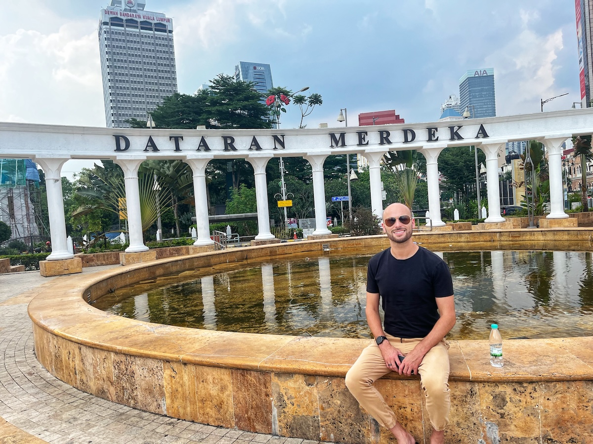 How Many Days Should You Spend in Kuala Lumpur? | Merdeka Square A man sitting on the edge of a circular fountain in front of the Dataran Merdeka sign in Kuala Lumpur, Malaysia, with tall buildings and lush greenery in the background