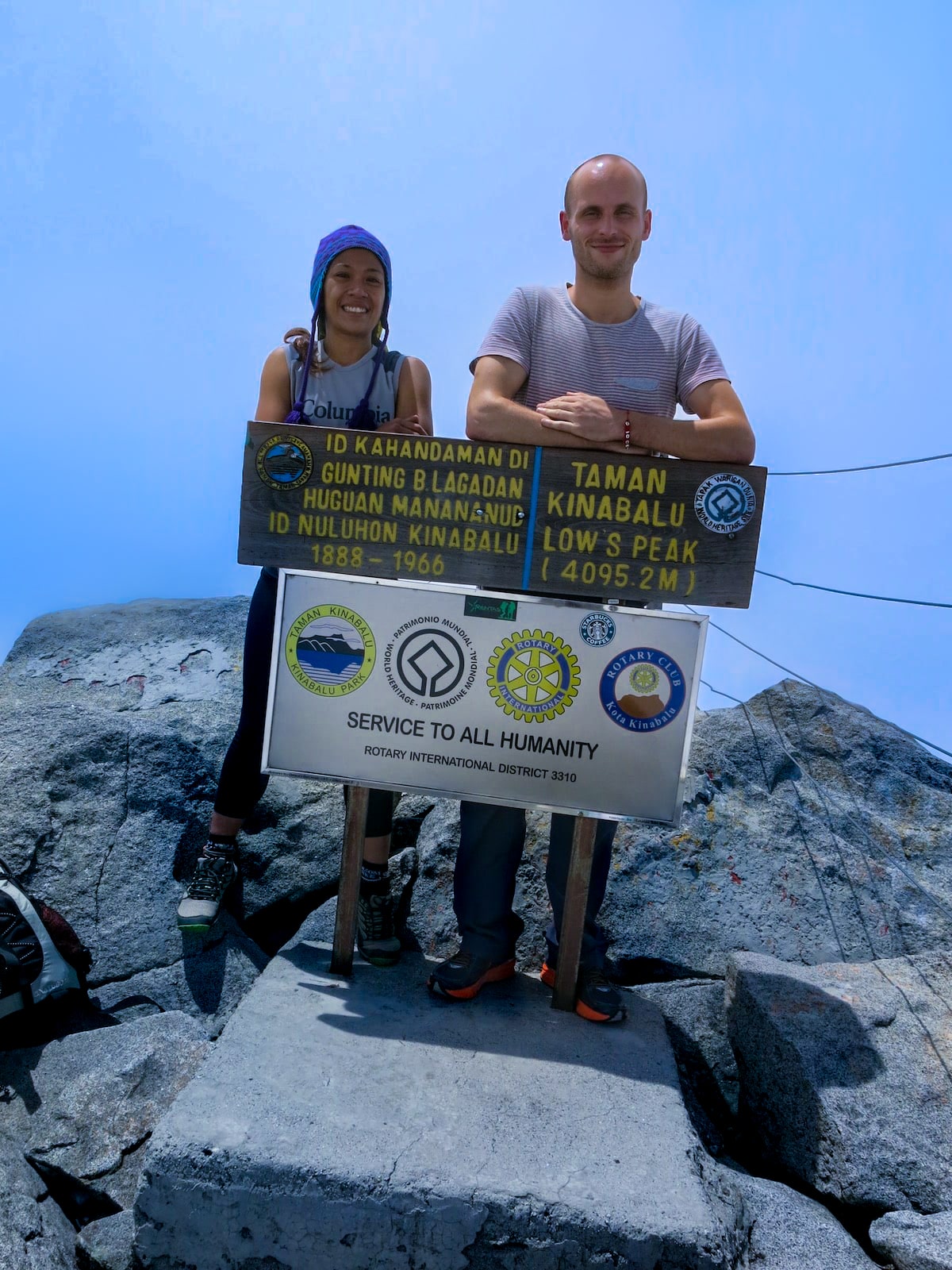 Climbing Mount Kinabalu Tips & Info For First-TimersĀ | Mount Kinabalu climb summit A man and a woman smile as they lean on a signpost on top of a mountain.