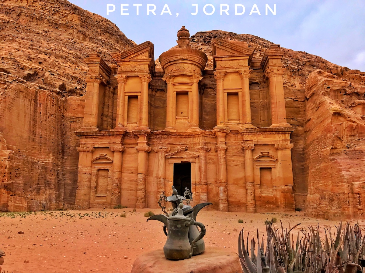 Ranking The New Seven Wonders Of The World From Worst To Best | Petra Jordan The Monastery (Al-Deir) in Petra, Jordan, a massive ancient building carved into sandstone cliffs, with a traditional teapot in the foreground, set against a backdrop of rugged desert landscape.