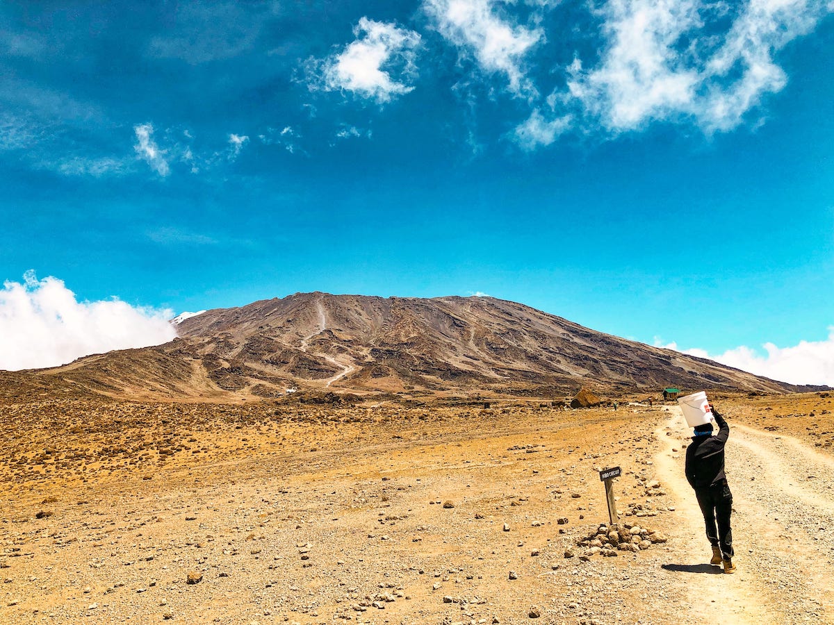 The Beginner's Guide To Climbing Mount Kilimanjaro | Porter carrying bucket on head A man walks towards a mountain with a bucket on his head.