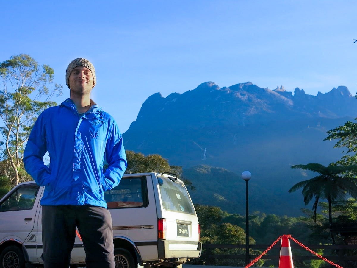 Climbing Mount Kinabalu Tips & Info For First-TimersĀ | Preparation for climbing Mount Kinabalu 2 A man in a grey hat and blue jacket poses at the bottom of a mountain.