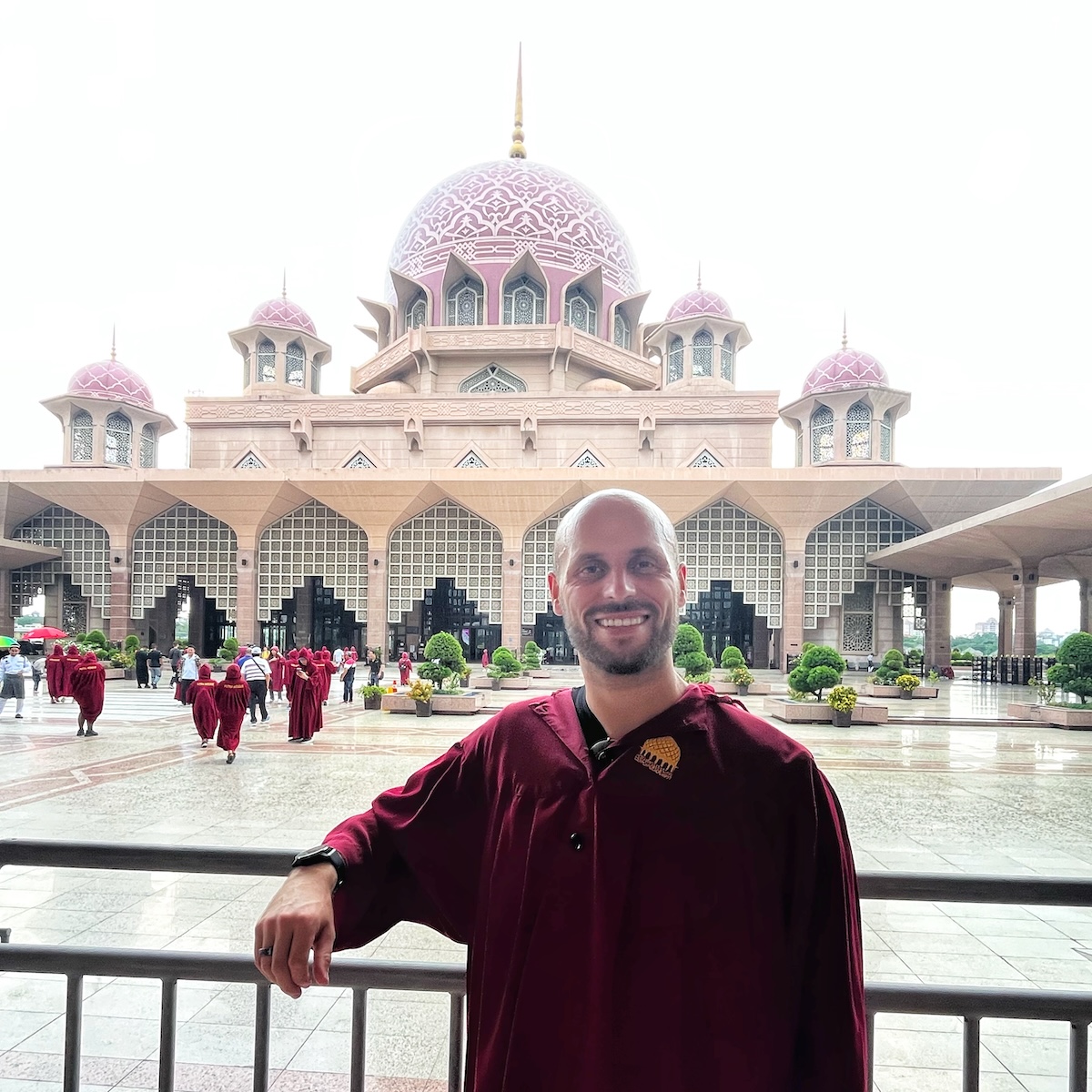 How Many Days Should You Spend in Kuala Lumpur? | Putra Mosque A man wearing a maroon robe standing in front of the Putra Mosque in Putrajaya, Malaysia, with its distinctive pink domes and intricate architectural design. Other visitors in similar robes can be seen in the background, highlighting the mosque's grandeur and cultural significance.