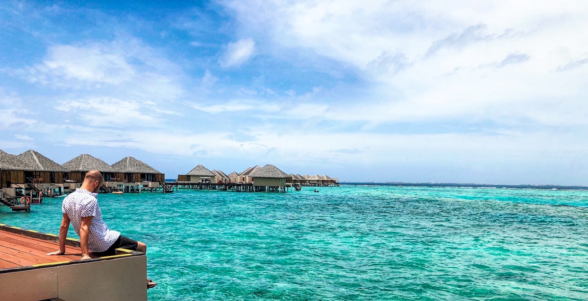 How To Plan a Trip To The Maldives | Tourist in Maldives A male tourist in shorts and shirt sits on the edge of a pier with the water bungalows behind him