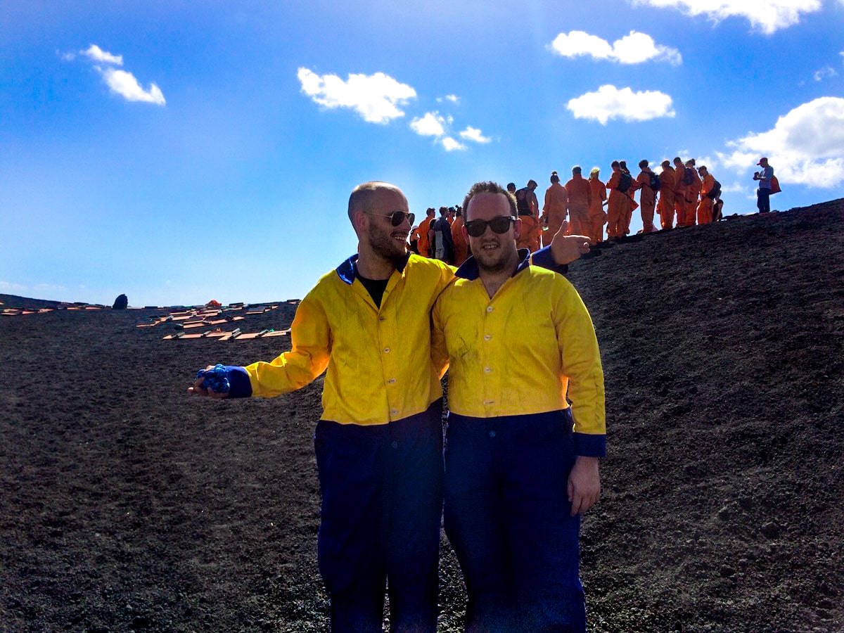 Volcano in Boarding in Leon, Nicaragua (Cerro Negro) | Volcano boarding clothes Two men in a yellow and blue jumpsuits stand on black volcanic ash in front of a tour group in orange jumpsuits.