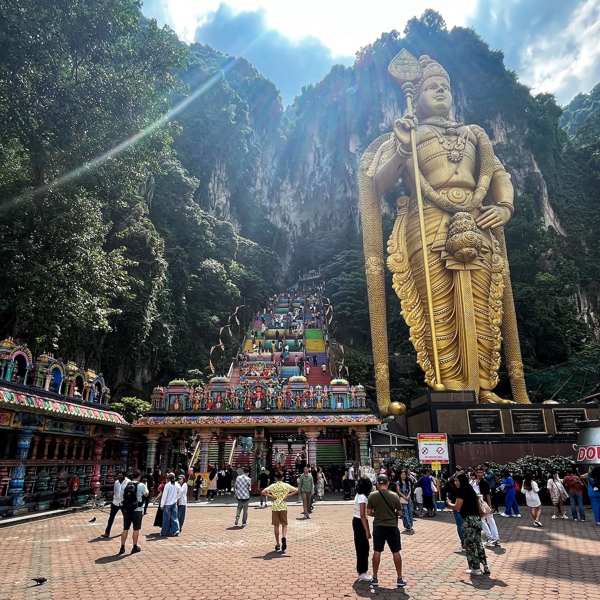 How Many Days Should You Spend in Kuala Lumpur? | batu caves lord murugan Visitors at the Batu Caves in Malaysia, with the towering golden statue of Lord Murugan and the vibrant, colorful staircase leading up to the caves set against a backdrop of lush green cliffs