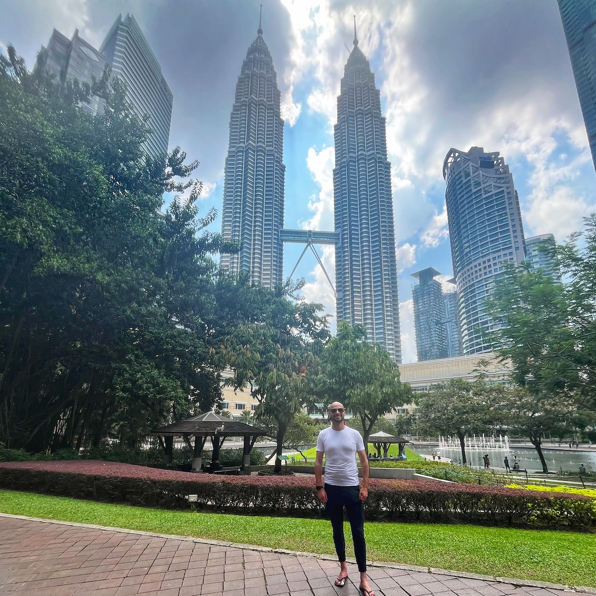 How Many Days Should You Spend in Kuala Lumpur? | best views Petronas Towers during day 1 A man standing in a park with the Petronas Twin Towers in Kuala Lumpur, Malaysia, towering in the background under a partly cloudy sky. The lush greenery of the park contrasts with the modern skyscrapers.