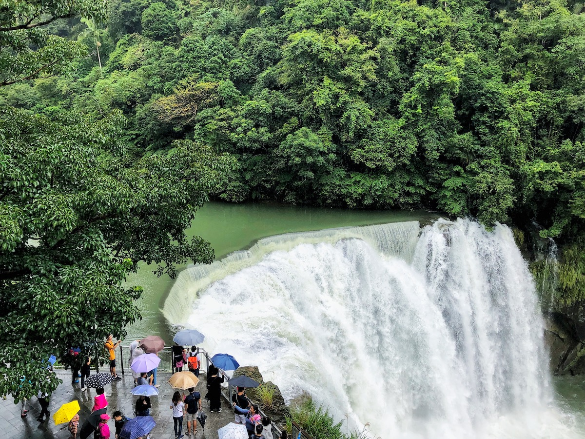 7 Unique & Fun Things To Do in Taipei (Guide For Weirdos!) | image 8 Tourists hold up their umbrellas near a large flowing waterfall under an emerald green lake and trees surrounding it