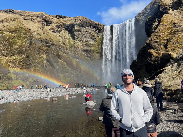 A male tourist smiles at a waterfall overlooking a rainbow in Skogafoss, Iceland