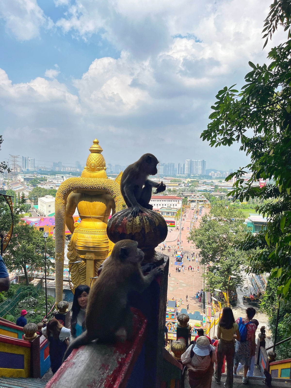 How Many Days Should You Spend in Kuala Lumpur? | monkeys at Batu Caves View from the Batu Caves in Malaysia, featuring two monkeys sitting on the colorful staircase railings with the golden statue of Lord Murugan and the cityscape in the background. Visitors are seen climbing the stairs, enhancing the lively atmosphere of the site.