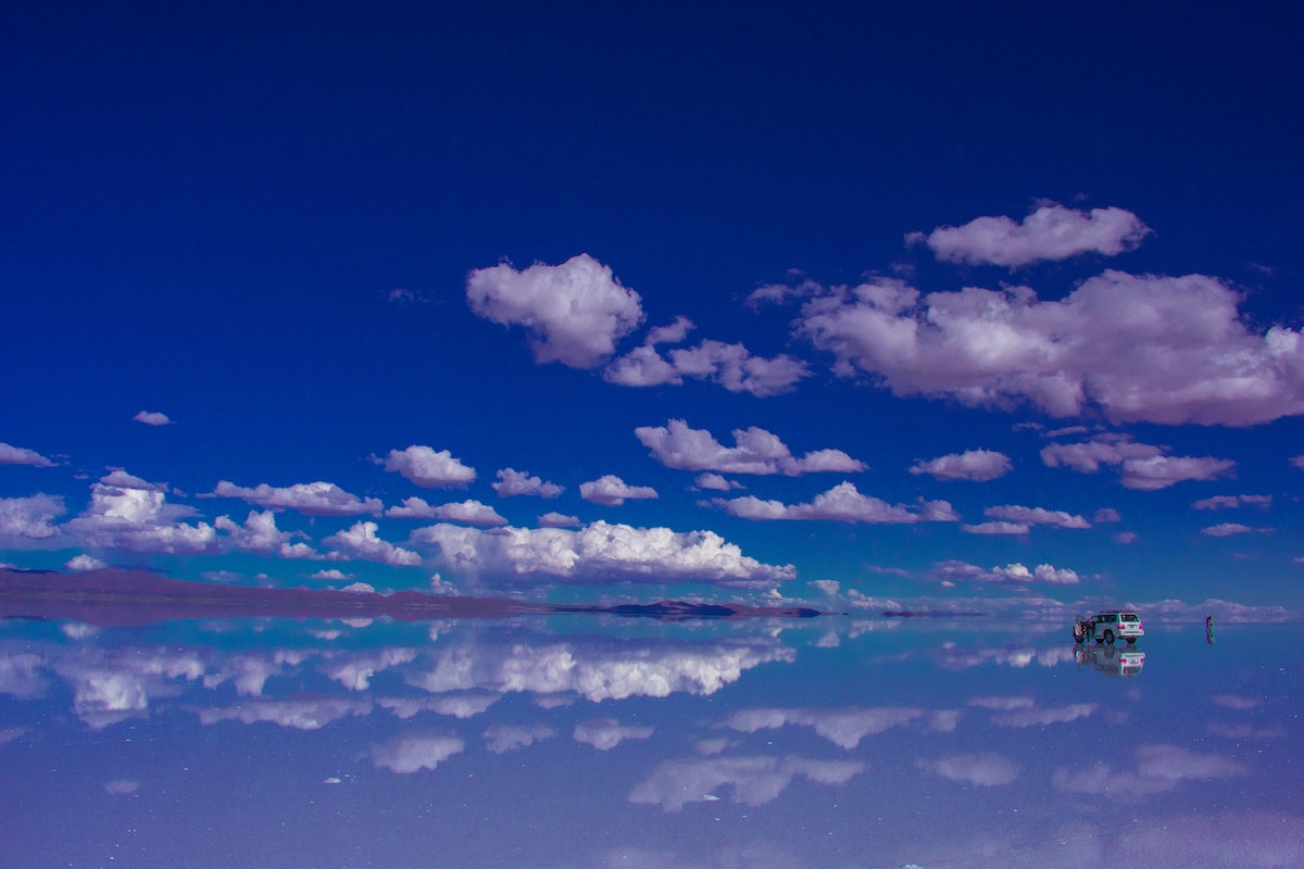 Visiting Salar de Uyuni in Bolivia: All You Need To Know | salar de uyuni mirror The sky reflects off the ground in a barren desert, people walk around a nearby jeep.
