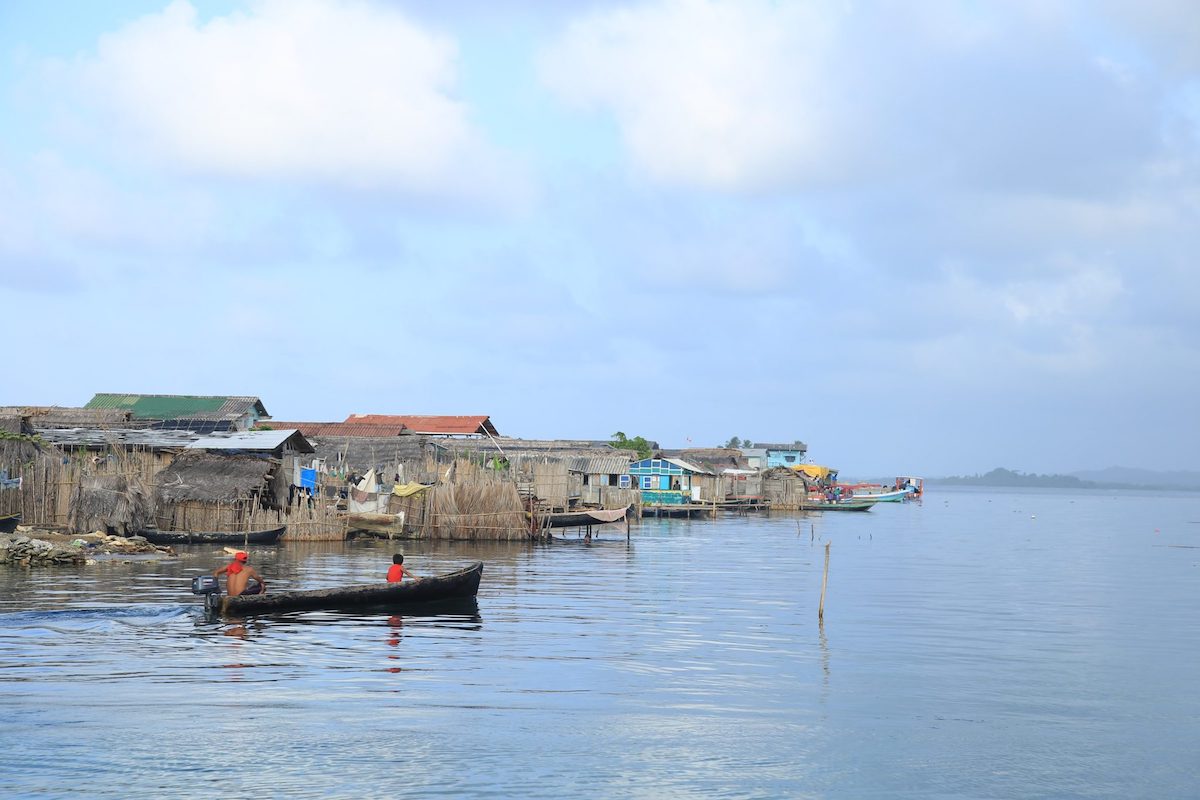 San Blas Islands Travel Guide: Best Things To Do & Logistics | san blas islands panama A man and a boy look onwards as they travel on a boat past wooden houses on stilts in the San Blas Islands, Panama.